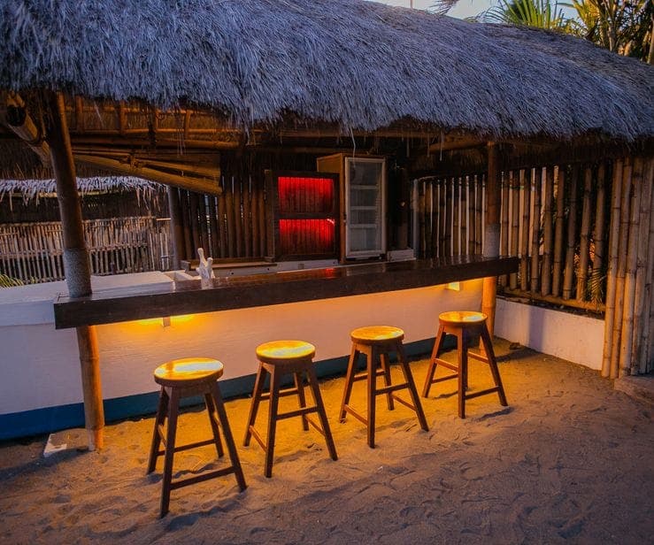 The beach bar at dusk, bamboo stools glowing under warm light