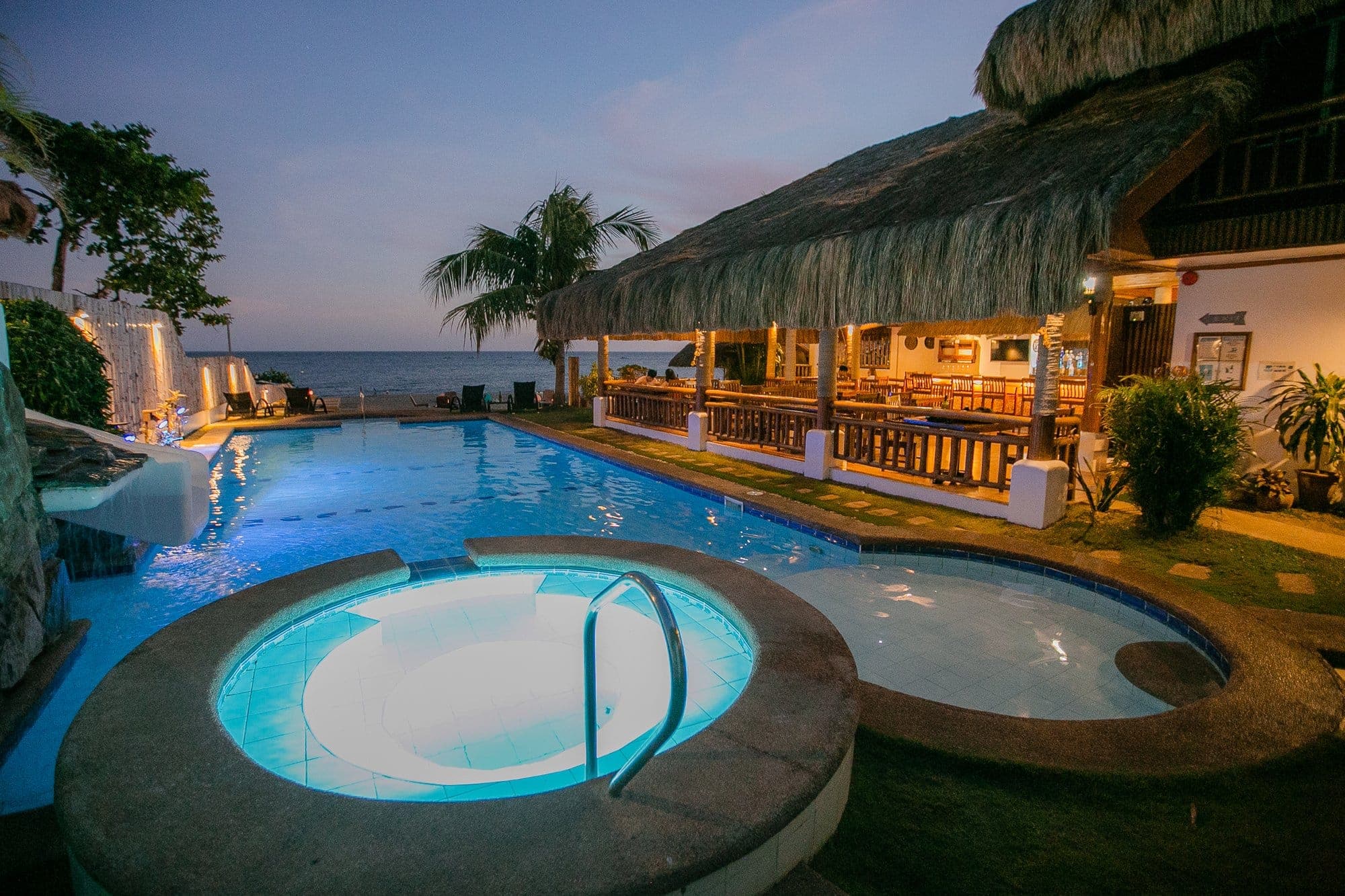 The resort pool at dusk with the thatched restaurant and sea beyond